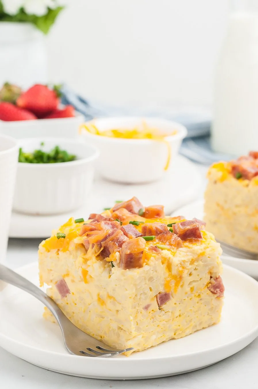 breakfast casserole serving on a plate alongside a fork. Strawberries, sliced green onions and cheddar cheese in individual bowls in the background.