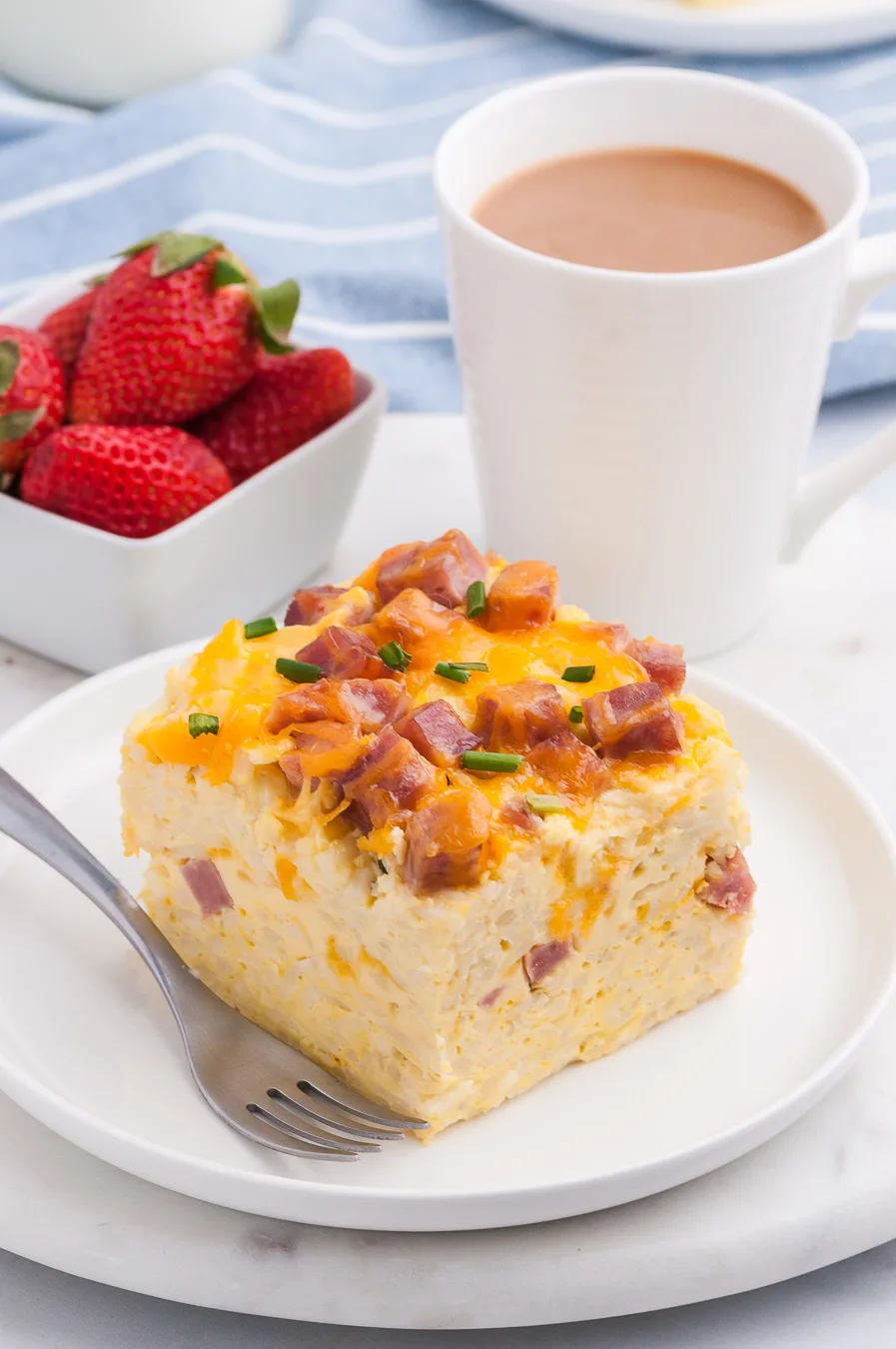 brunch casserole with ham and cheddar cheese made in the slow cooker. Serving placed on a plate and served next to coffee mug and fresh strawberries.