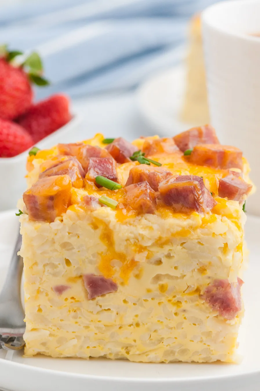 hearty slice of breakfast casserole sliced into a square portion. Strawberries and coffee mug in background.