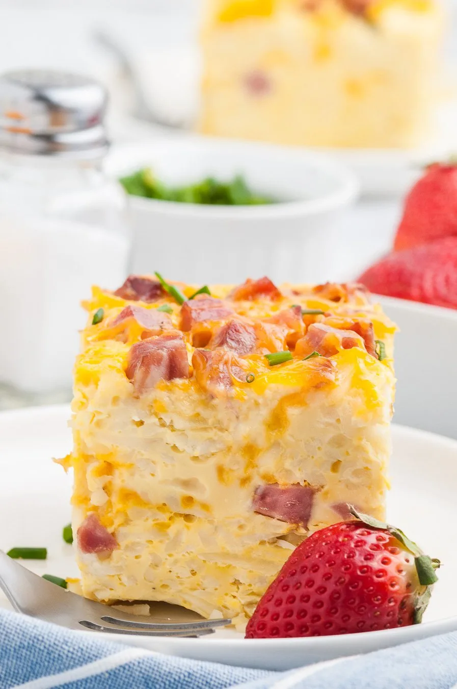 tall breakfast casserole on a dish with chunks of ham showing through. Paired with a fresh strawberry garnish. Hint of blue napkin near plate.