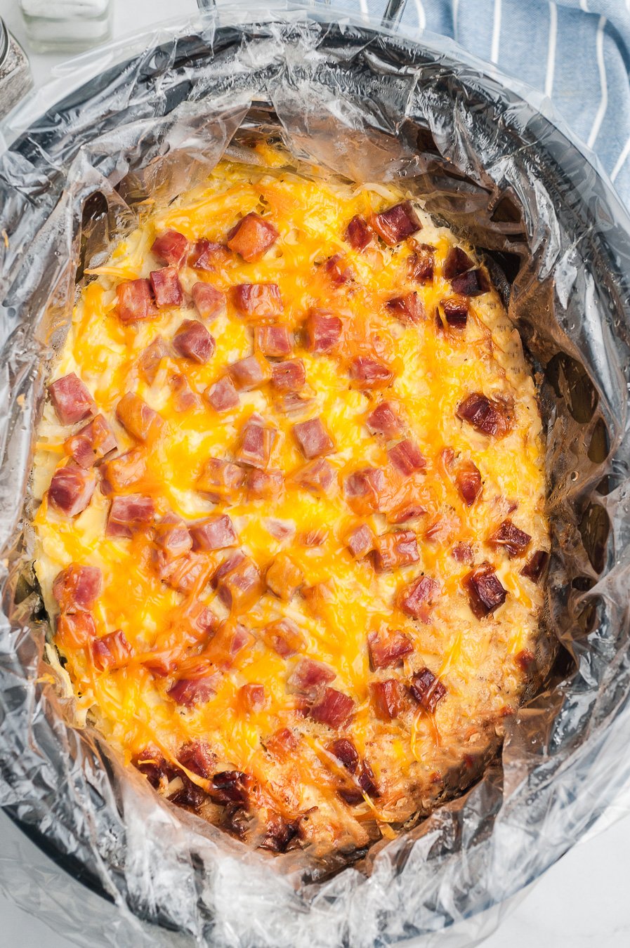 inside view of slow cooker with prepared breakfast casserole inside with chunks of cooked ham and melted cheddar cheese.