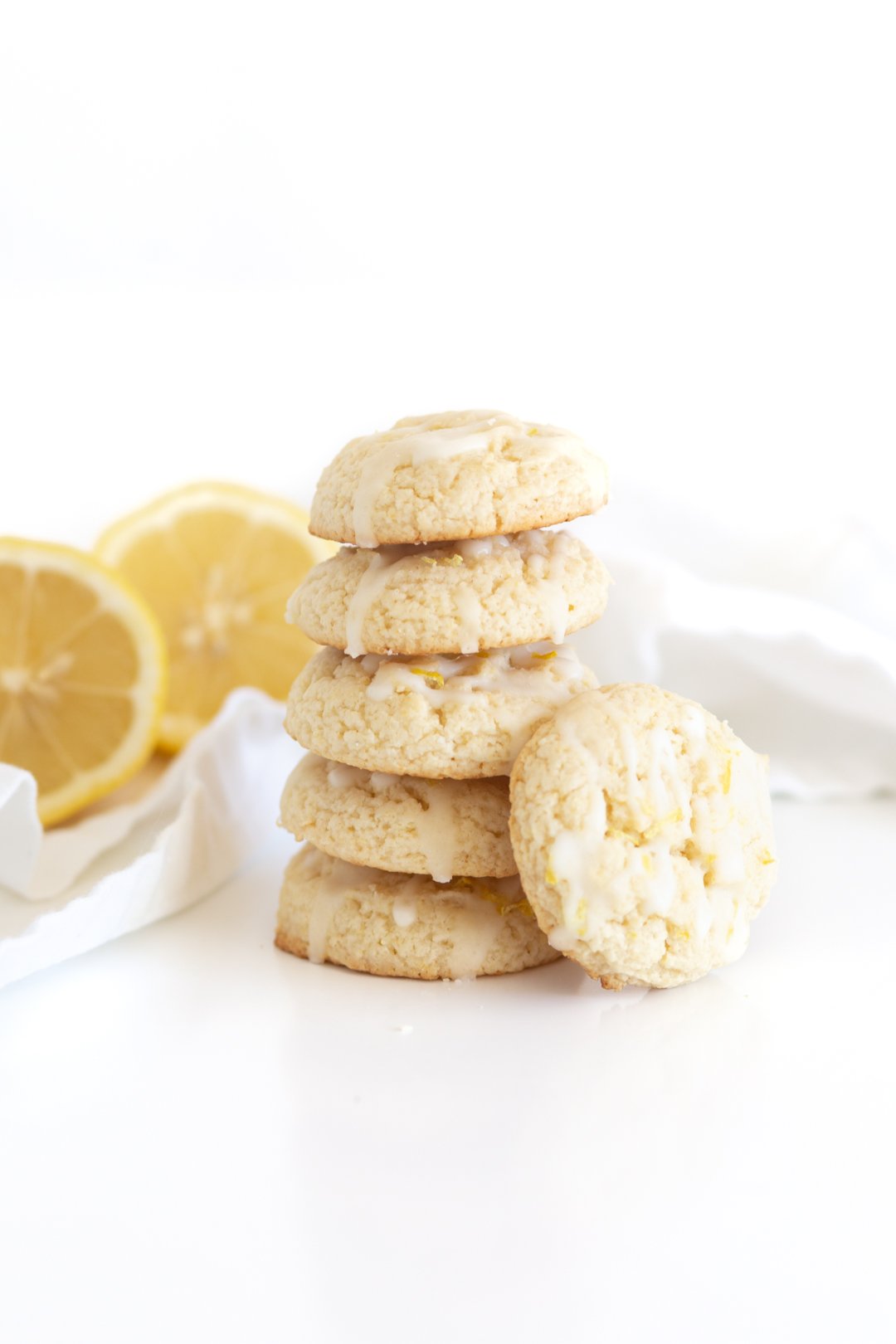 stack of five lemon cookies with an additional cookie leaning on the stalk. Halved lemons in the background.