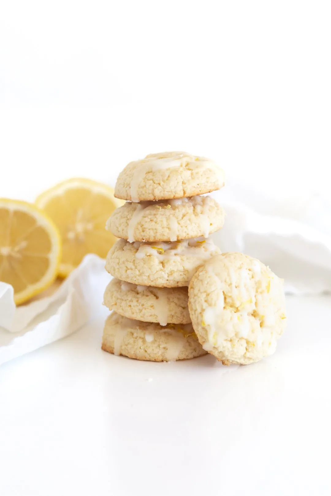 stack of five lemon cookies with an additional cookie leaning on the stalk. Halved lemons in the background.