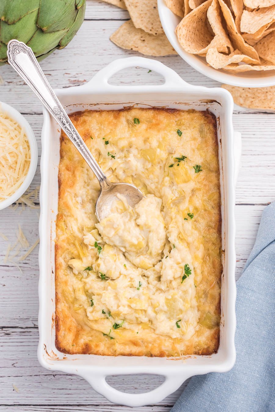 overhead photo of a baking dish filled with artichoke dip with curly parsley garnish. Tortilla chips on the side.