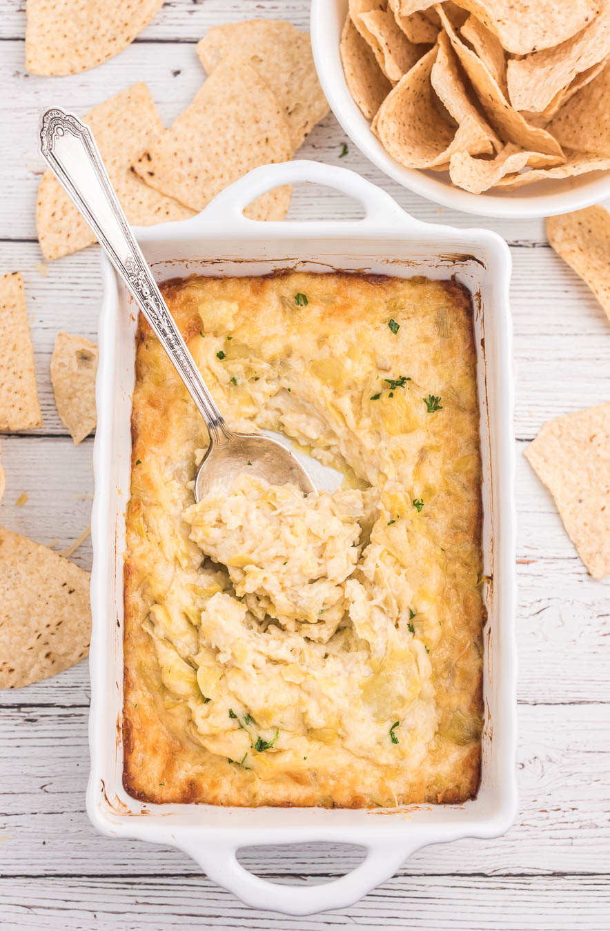 artichoke dip served in a white baking dip with a spoon. Tortilla chips on the side.