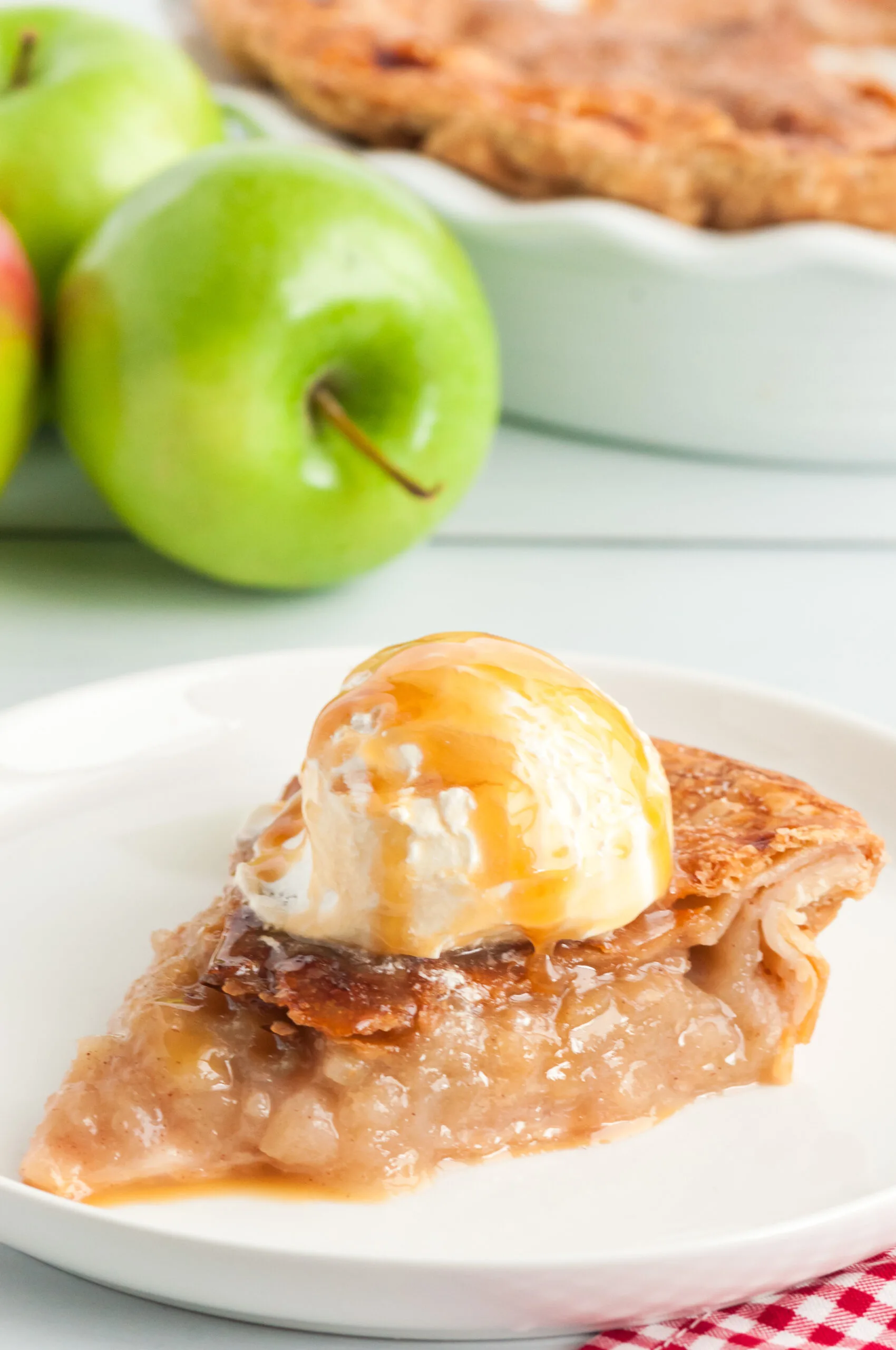 single slice of hot apple pie with ice cream and caramel drizzle on top. fresh apples in background.