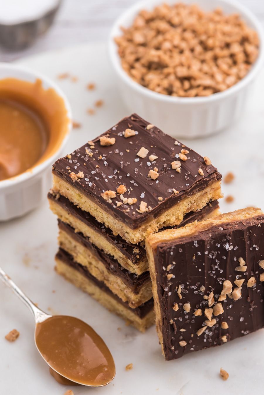 stack of chocolate covered cookie bars surrounded by a spoonful of caramel, mini dishes of caramel and toffee bits
