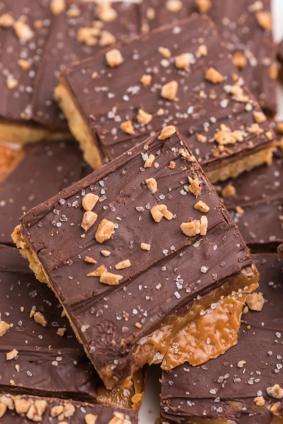 up close of a stack of chocolate covered cookie bars topped with sea salt and toffee bits with caramel oozing out