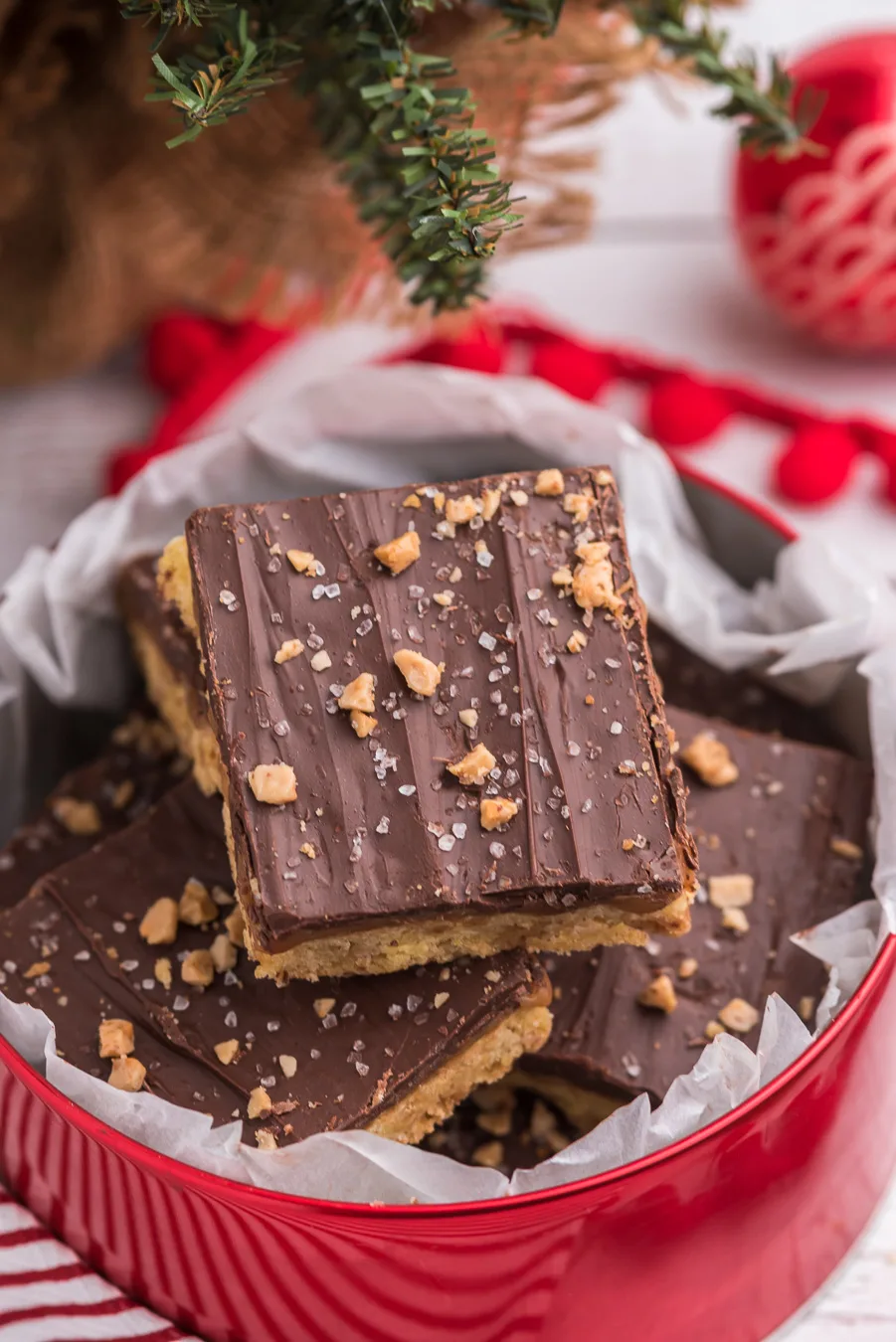chocolate covered caramel bars topped with toffee bits and sea salt. bar stacked into a red Christmas tin. table top Christmas tree and red ornament in background
