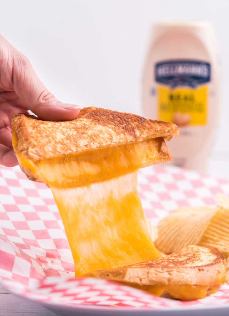 amazing cheese pull from a simple grilled cheese sandwich being held by a woman's hand. Bottle of hellmann's in the background.