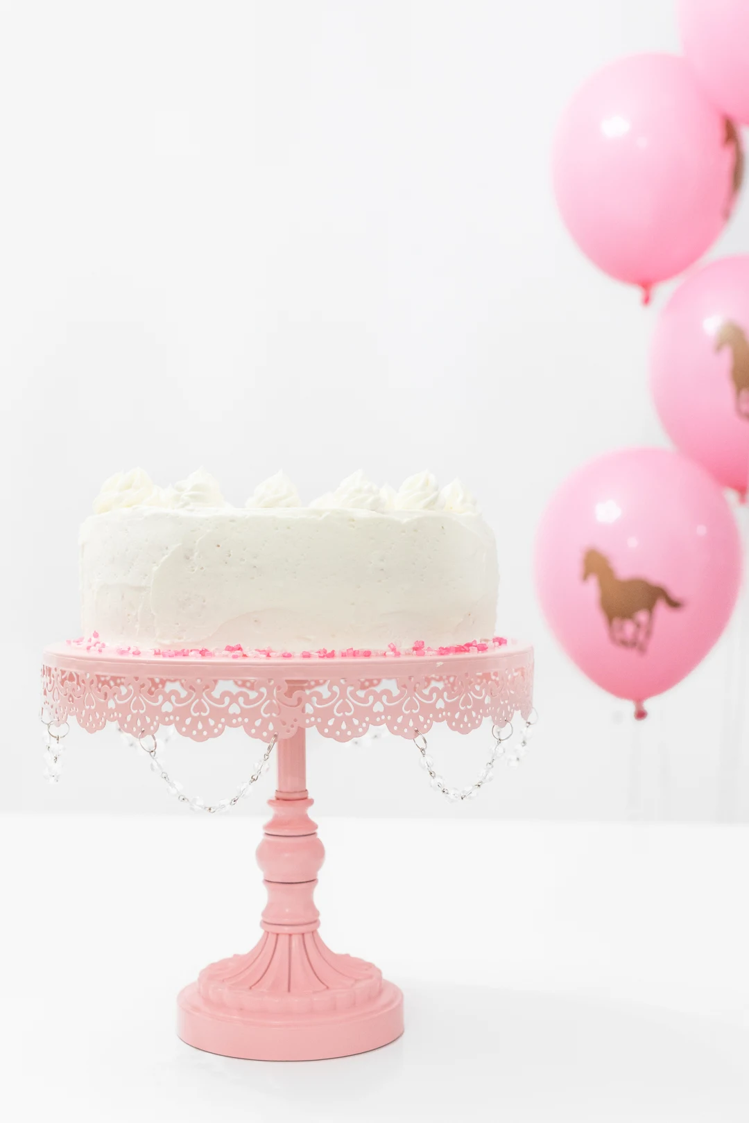 White cake on pretty pink cake stand. Horse balloons.