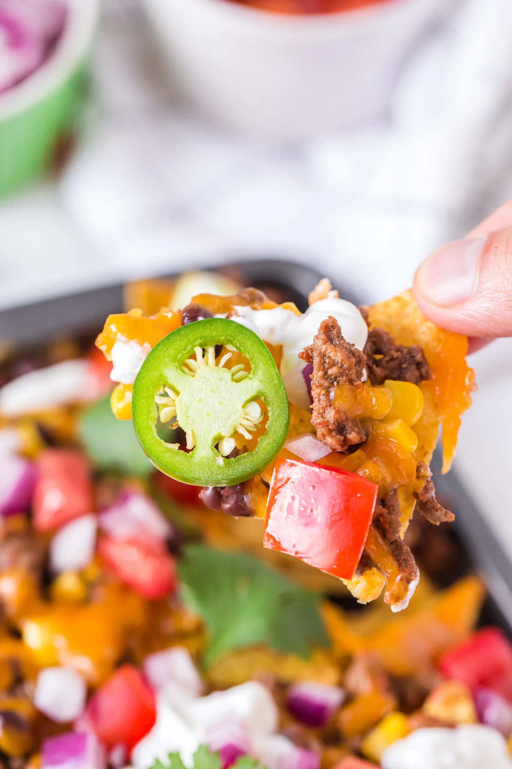 woman holding a single nacho loaded with tomatoes, cheese, beef, sour cream and sliced jalapeños