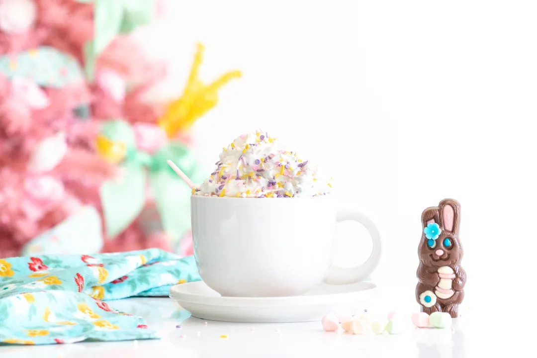 easter bunny hot cocoa melt near a mug filled with whipped cream and topped with sprinkles. pink easter christmas tree in the background with pastel decorations on it.