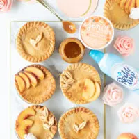 tray of mini peach pies with embellishments on top, spoonful of caramel, can of whipped cream, ice cream. Faux peach flowers for decoration and peach paper banner.