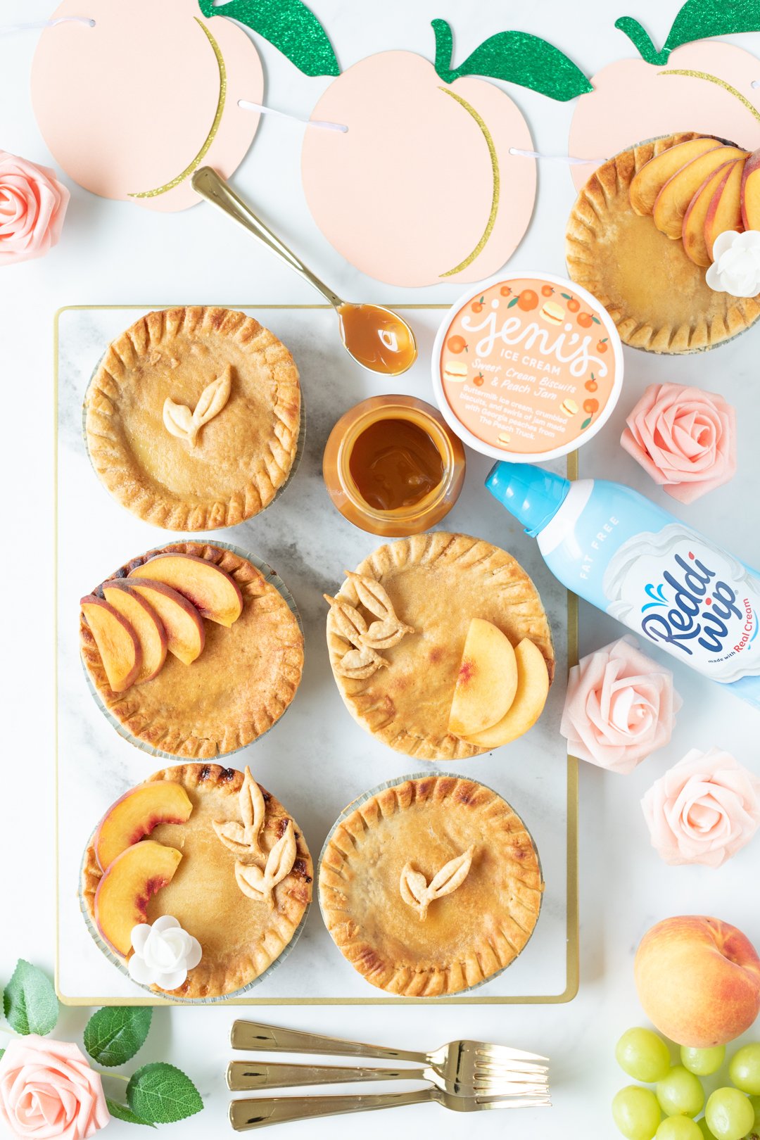 tray of mini peach pies with embellishments on top, spoonful of caramel, can of whipped cream, ice cream. Faux peach flowers for decoration and peach paper banner.