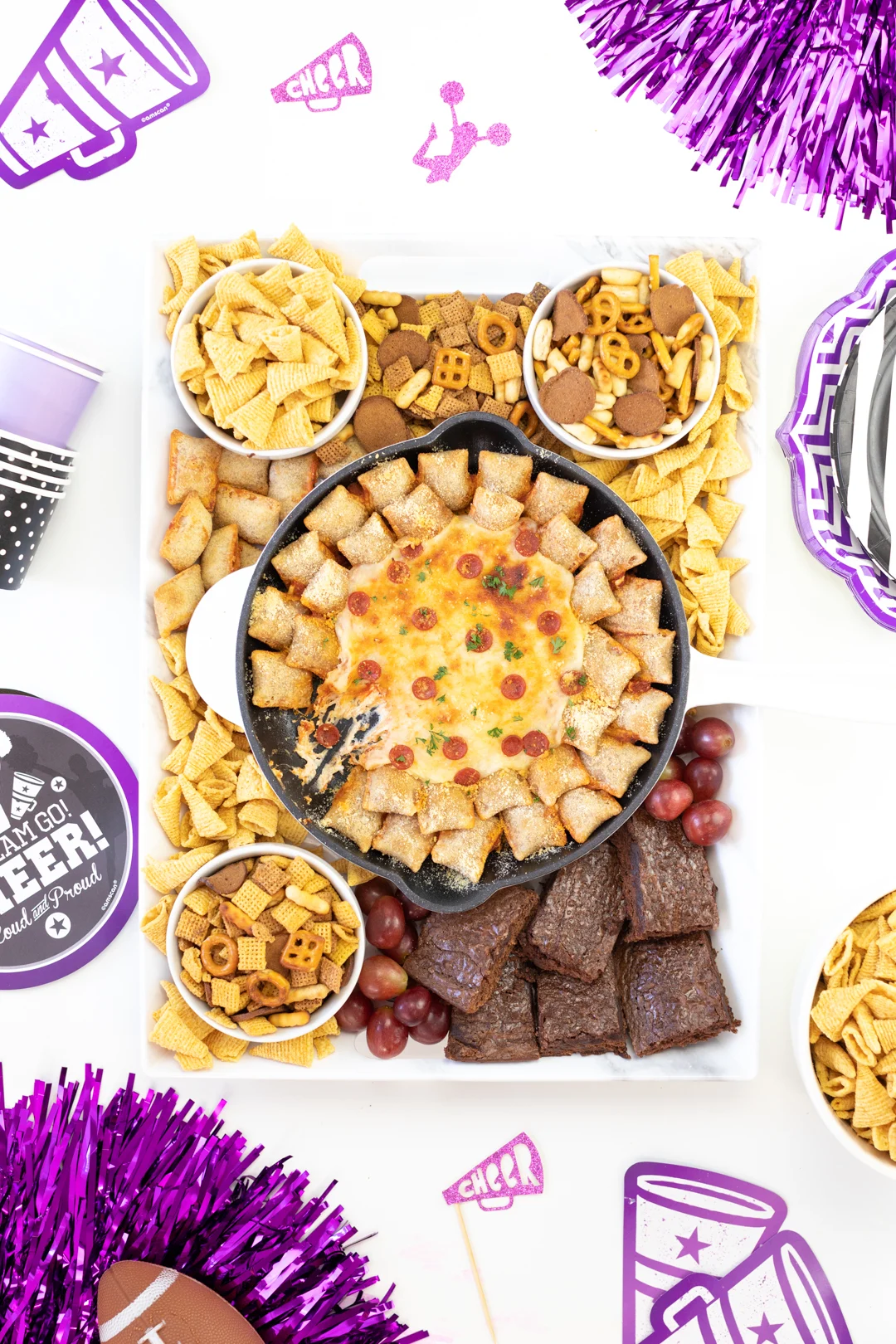 pretty tray of football snacks with pepperoni skillet dip, chex mix, bugles, brownies, grapes. Tablescape decorated with purple and black cheerleading party supplies. Alternative for football decorations.