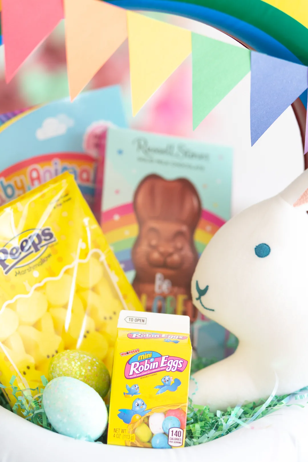 up close view of easter basket fillers including robin's eggs, yellow peeps bunnies, chocolate bunny and stuffed bunny pillow. Rainbow pendent banner made with construction paper.