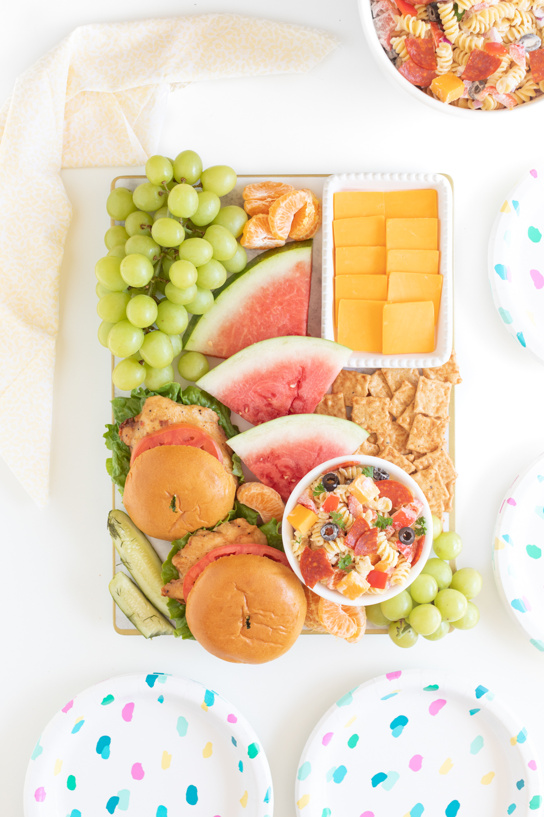 bbqterie board with grilled chicken sandwiches on buns, sliced watermelon, fruits, pasta salad, cheese and crackers