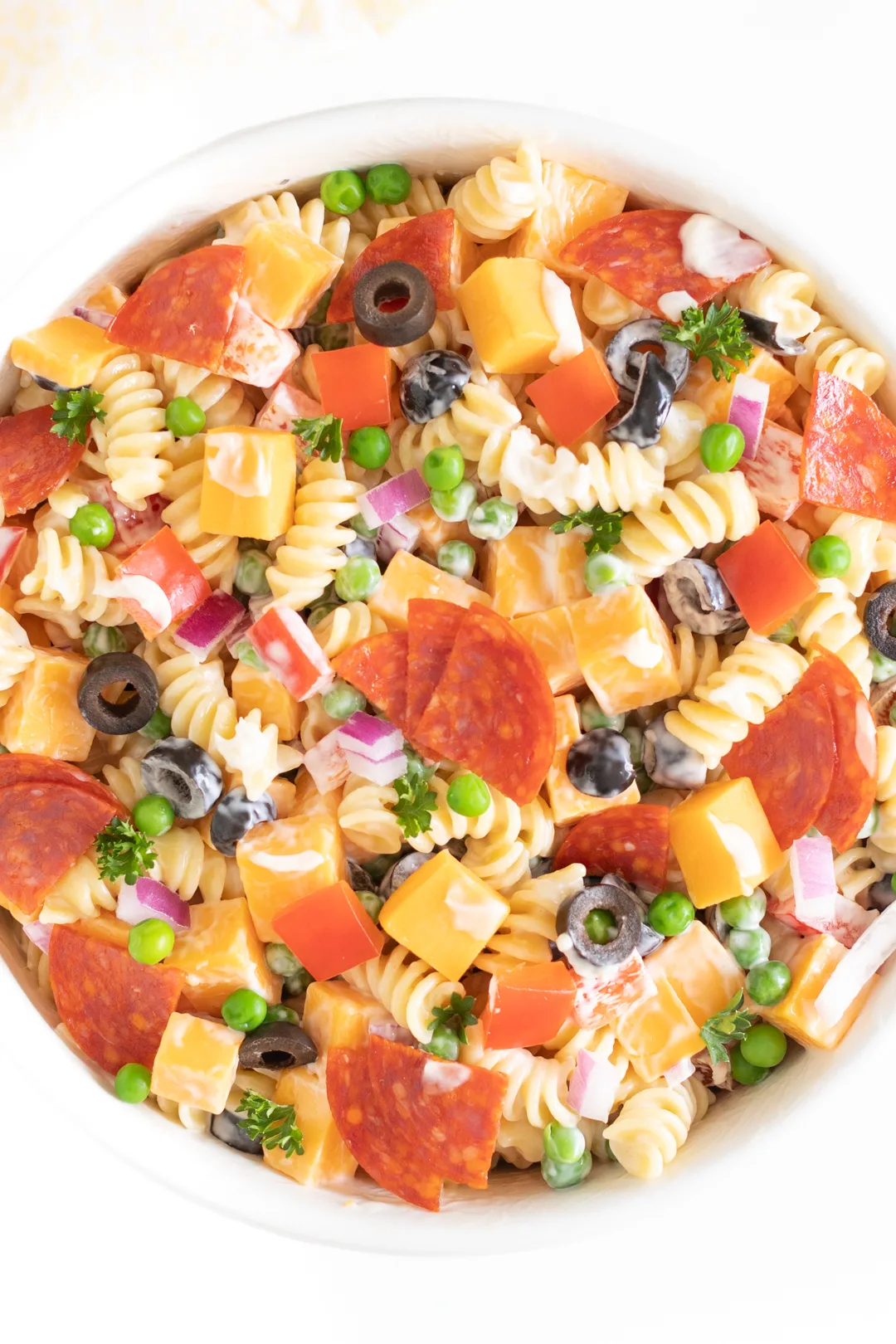 up close of pasta salad in a bowl topped with cheese chunks, pepperoni quarters, parsley, chopped red peppers and more.