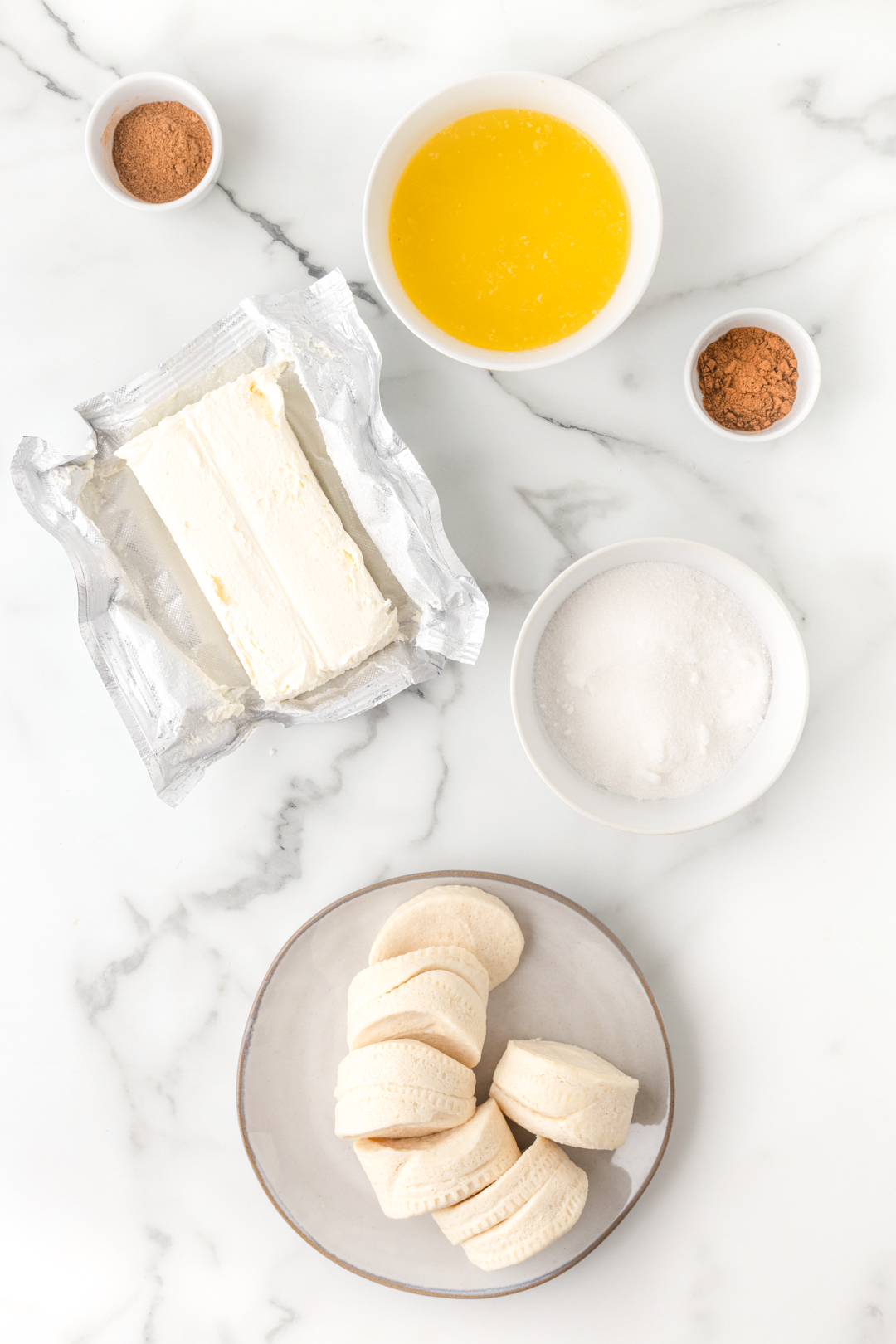 ingredients needed to make pumpkin spice donut holes in the air fryer. Canned biscuits on a plate, sugar, melted butter, cream cheese, cinnamon. 