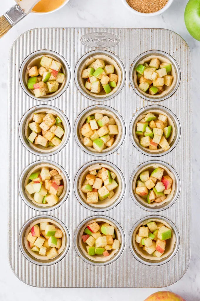 preparing mini apple pies in a muffin tin, chopped apples placed on top of dough circles