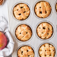 close up of individual apple pies baked in a muffin tin with fresh apples surrounding it