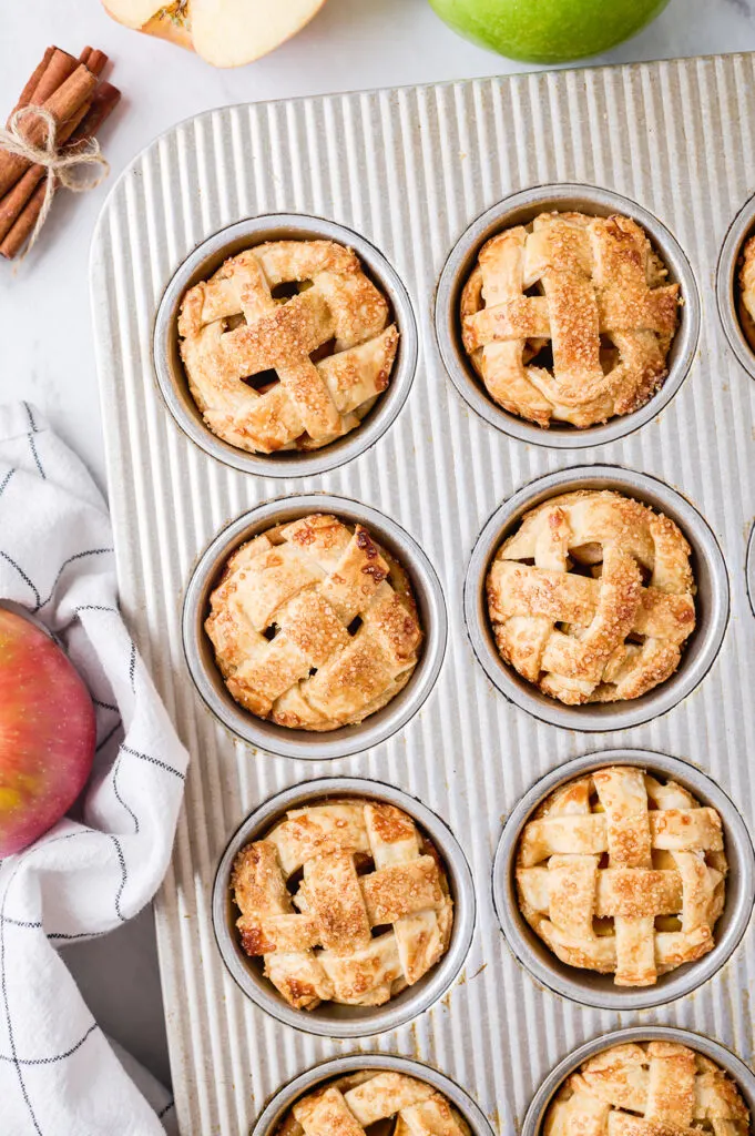 close up of individual apple pies baked in a muffin tin with fresh apples surrounding it