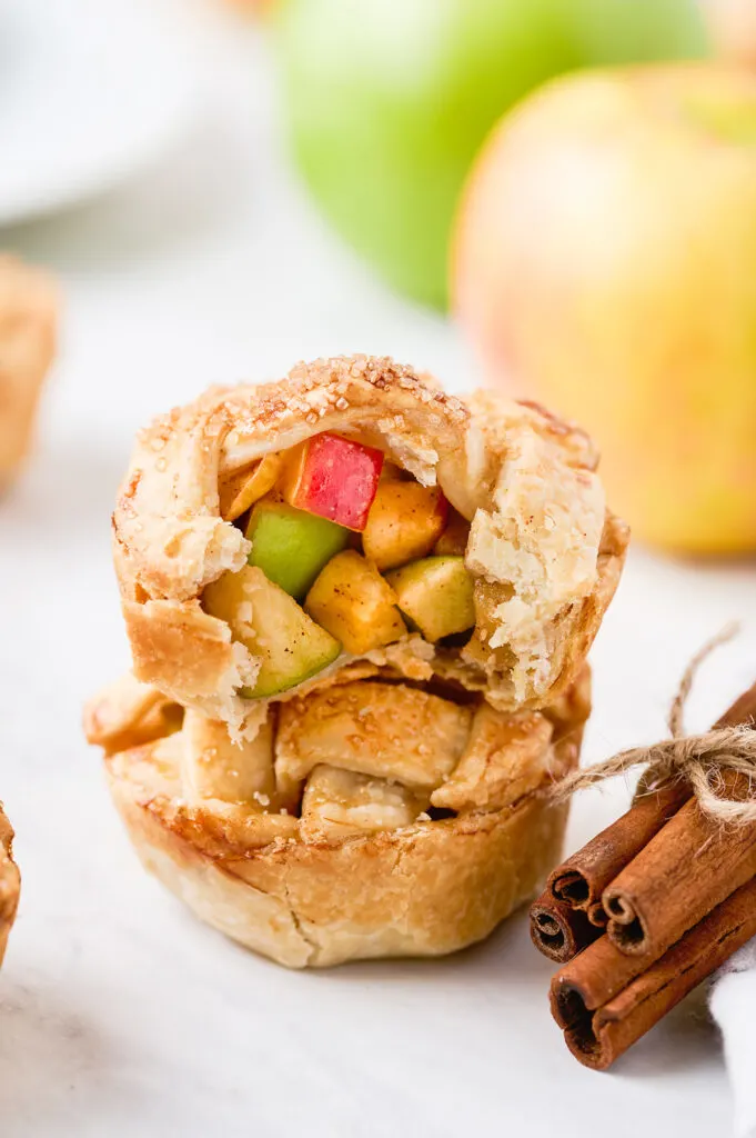 two mini apple pies stacked on top of each other. the top pie has a bite taken out of it to reveal the pretty red and green apple chunks inside