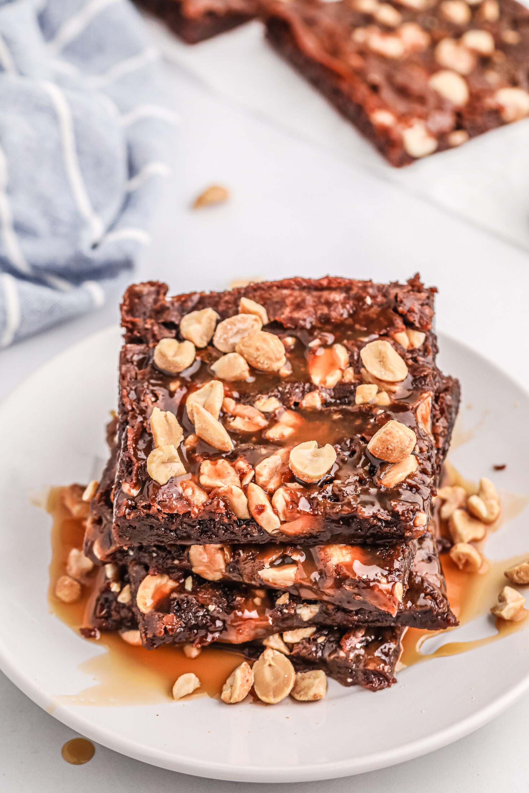 close view of thin brownies stacked on a small white plate and topped with a lot of roasted peanuts and caramel drizzle