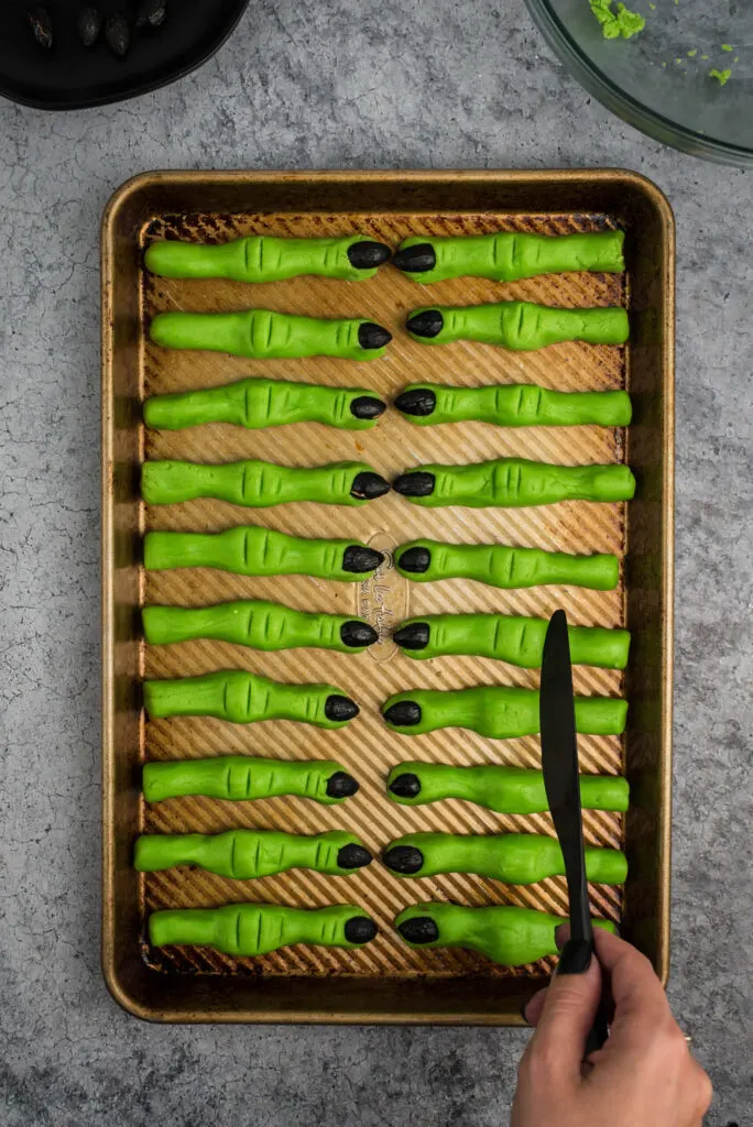 Green witch cookies with black fingernails on a baking sheet. Woman's hand holding a black plastic knife to show how to make indents into the cookies to make them look like fingers