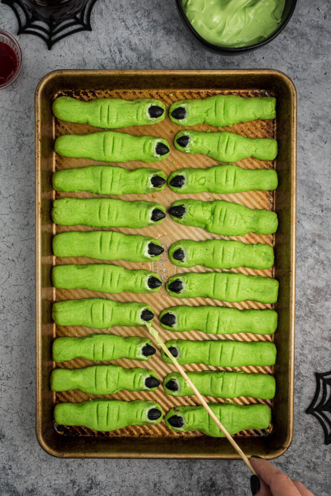 over the top photo of witch finger cookies on a baking sheet. Using a wooden skewer to finish decorating