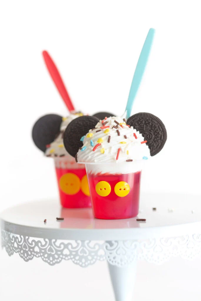 mickey mouse themed gelatin cups using snack packs with yellow buttons attached and topped with whipped cream, mickey mouse themed sprinkles, oreos for ears and red and blue spoons