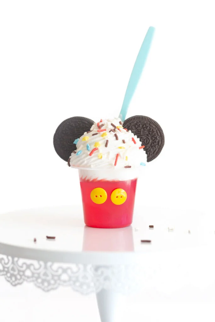 Mickey mouse jello cup decorated with yellow buttons, oreos, whipped cream and mickey mouse colored sprinkles
