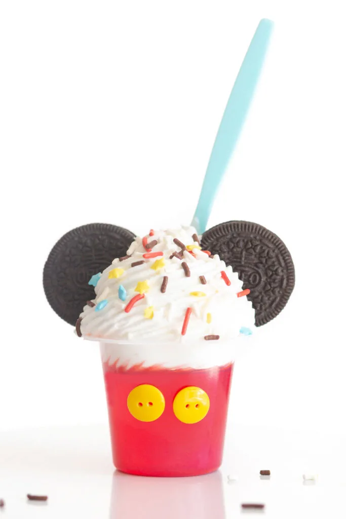 up close view of mickey mouse party dessert cup decorated to look like mickey mouse with yellow buttons, oreo cookies, whipped cream and matching colored sprinkles