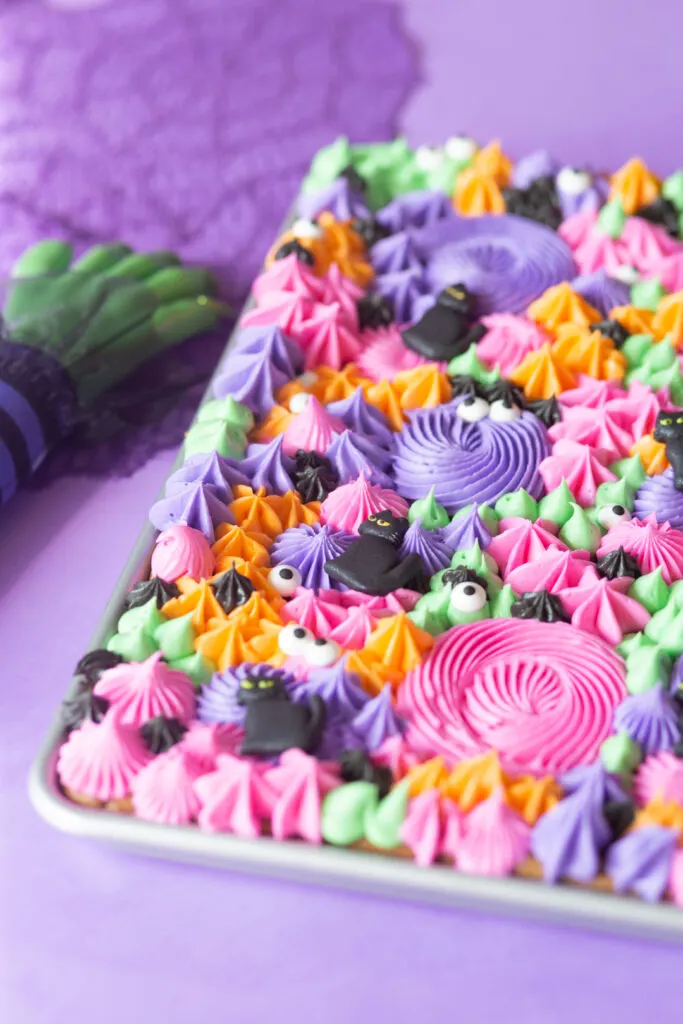 angled shot of buttercream cookie sheet with pink, green, orange, purple and black. Witch hand prop next to it.