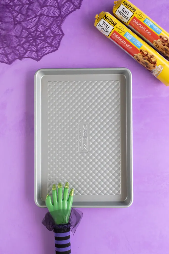 half sheet pan and two rolls of nestle cookie dough rollls on a purple backdrop. Purple web table runner in the corner. Witch hand prop resting on the sheet pan.