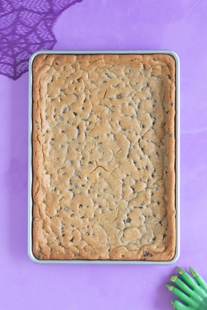 baked cookie dough on a half baking sheet on a purple backdrop. Purple web decoration in top left corner. Witch hand in bottom right corner.