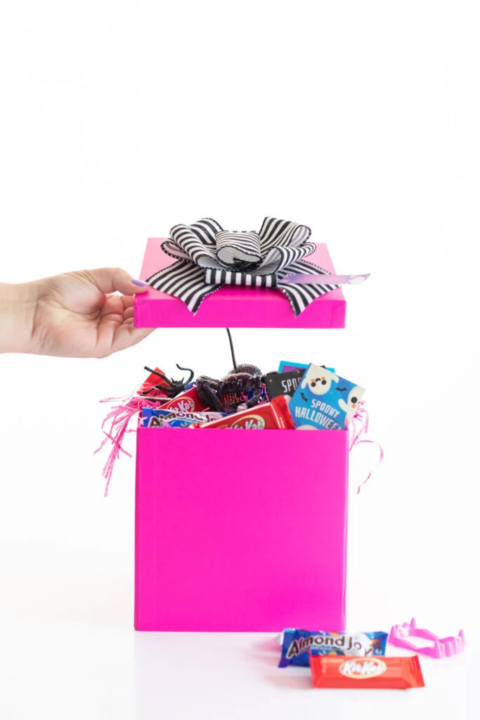 taking the top off of a gift box to reveal a spooky surprise for halloween. pretty bright pink box with a black and white striped bow on top