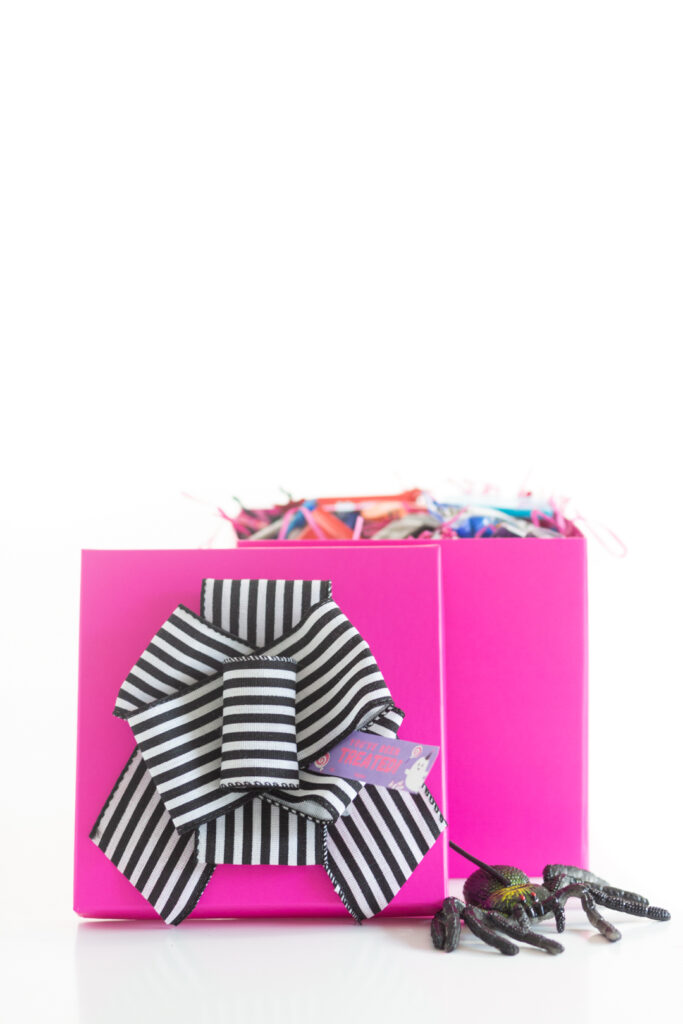 bright pink halloween gift boxes with thick black and white striped bow on top with a rubber spider near by to spook gift recipient