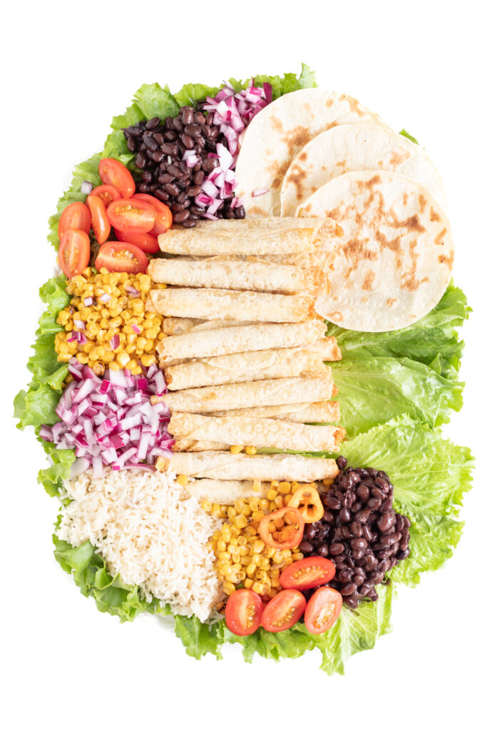 taquito platter being made over a bed of lettuce on a large serving tray. Ingredients stacked over the lettuce.