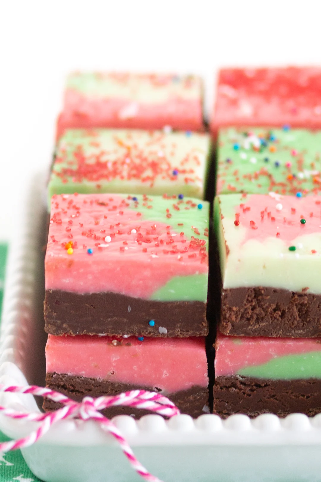 super close view of a stack of christmas fudge. two layered fudge. bottom layer, all chocolate. top layer white chocolate tinted red and green and swirled. Topped with variety of sprinkles.