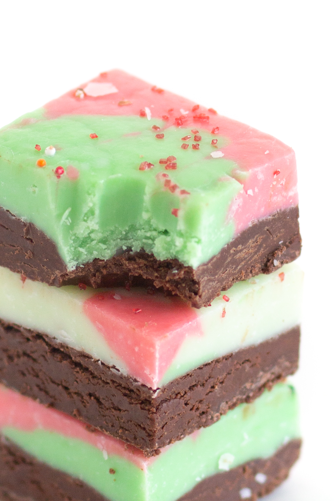 squares of multi colored fudge stacked on top of each other. Top square with a bite taken out if it. toped with red crystal sprinkles.