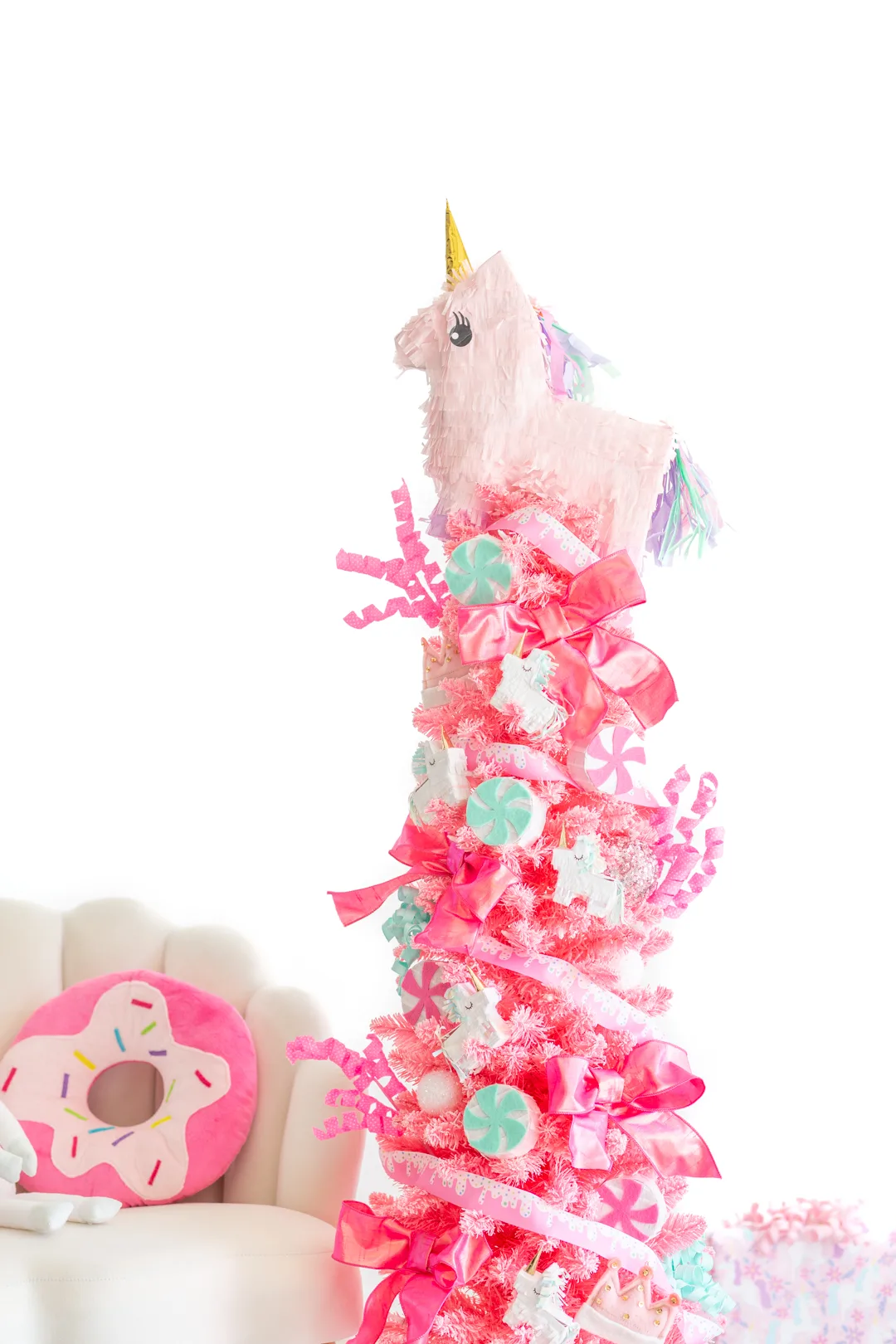 thin pink christmas tree decorated with a unicorn theme. next to a cream colored shell chair with a donut pillow resting on it