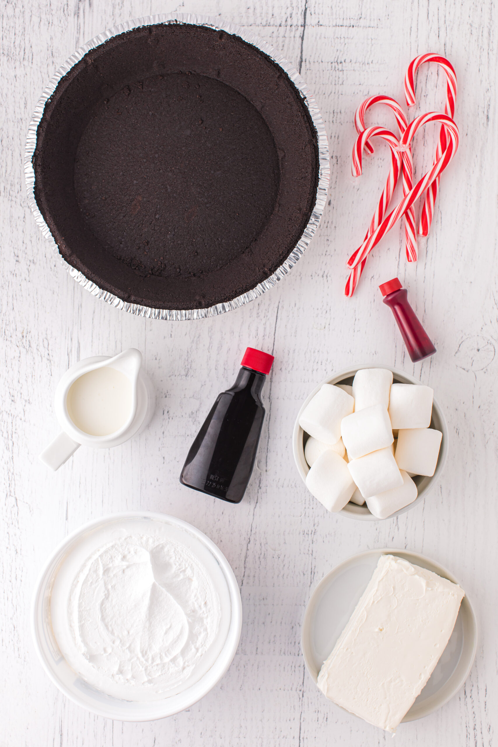 ingredients to make a no bake layered peppermint pie. oreo pie crust, peppermint extract, whipped topping, brick of cream cheese, large marshmallows, red and white striped candy canes