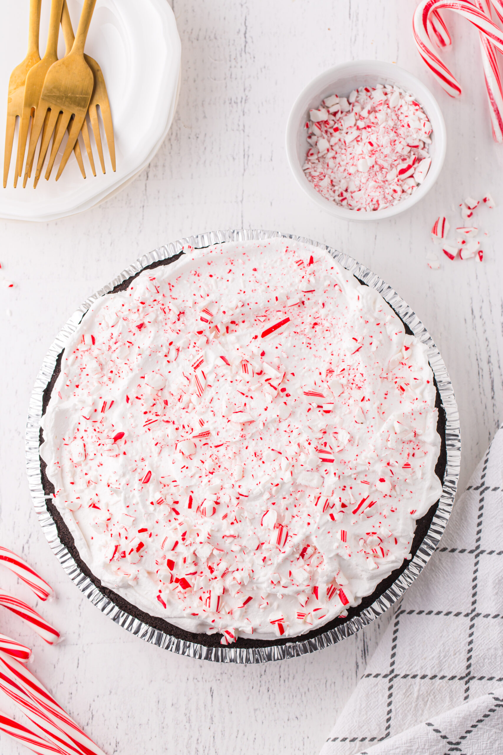 peppermint pie with whipped topping and crushed peppermint sprinkled over the top. small bowl of crushed peppermint, small plate with fold forks on top, whole candy canes and cloth napkin surrounding it.