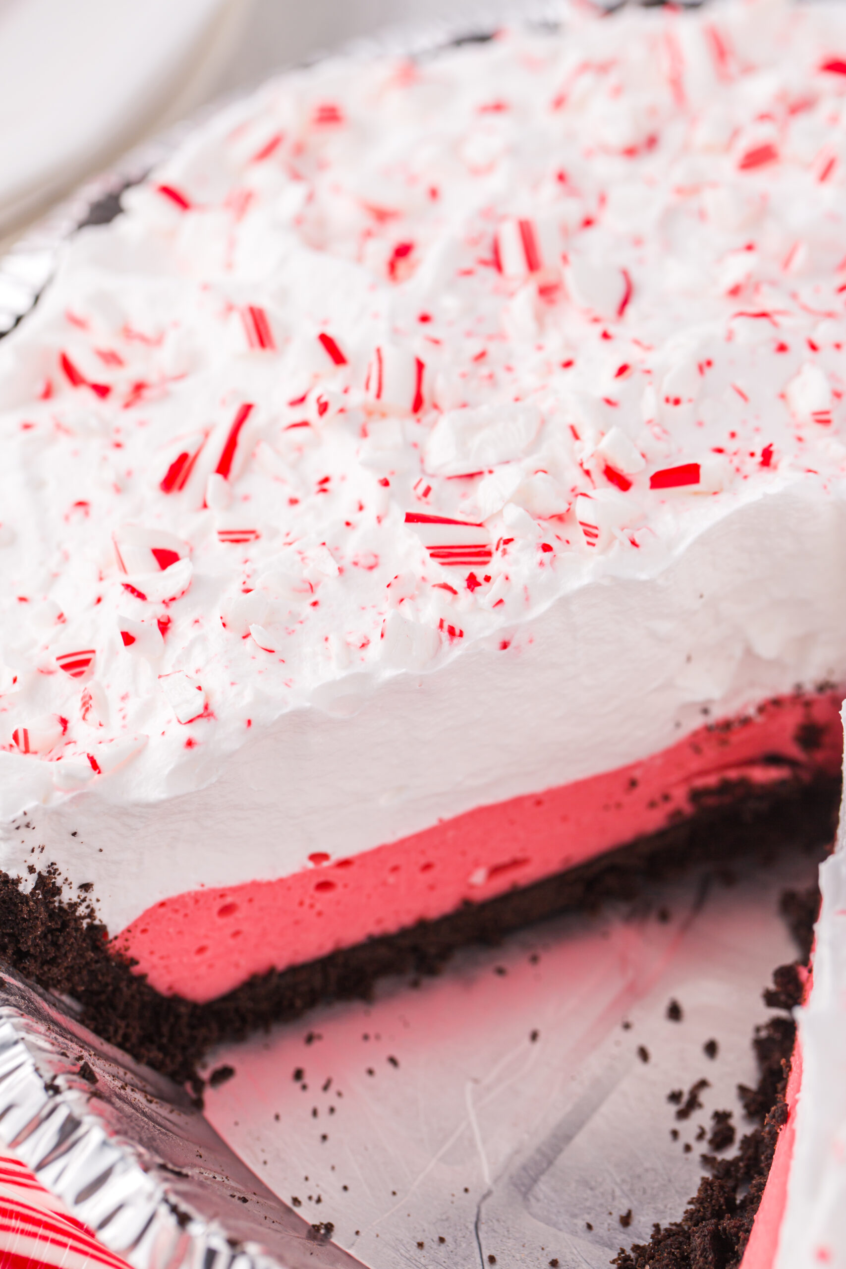 angled shot of peppermint pie with a slice missing. Pretty layers of crushed oreo crust, red peppermint marshmallow layer and then topped with whipped topping layer and toped with crushed red and white peppermint