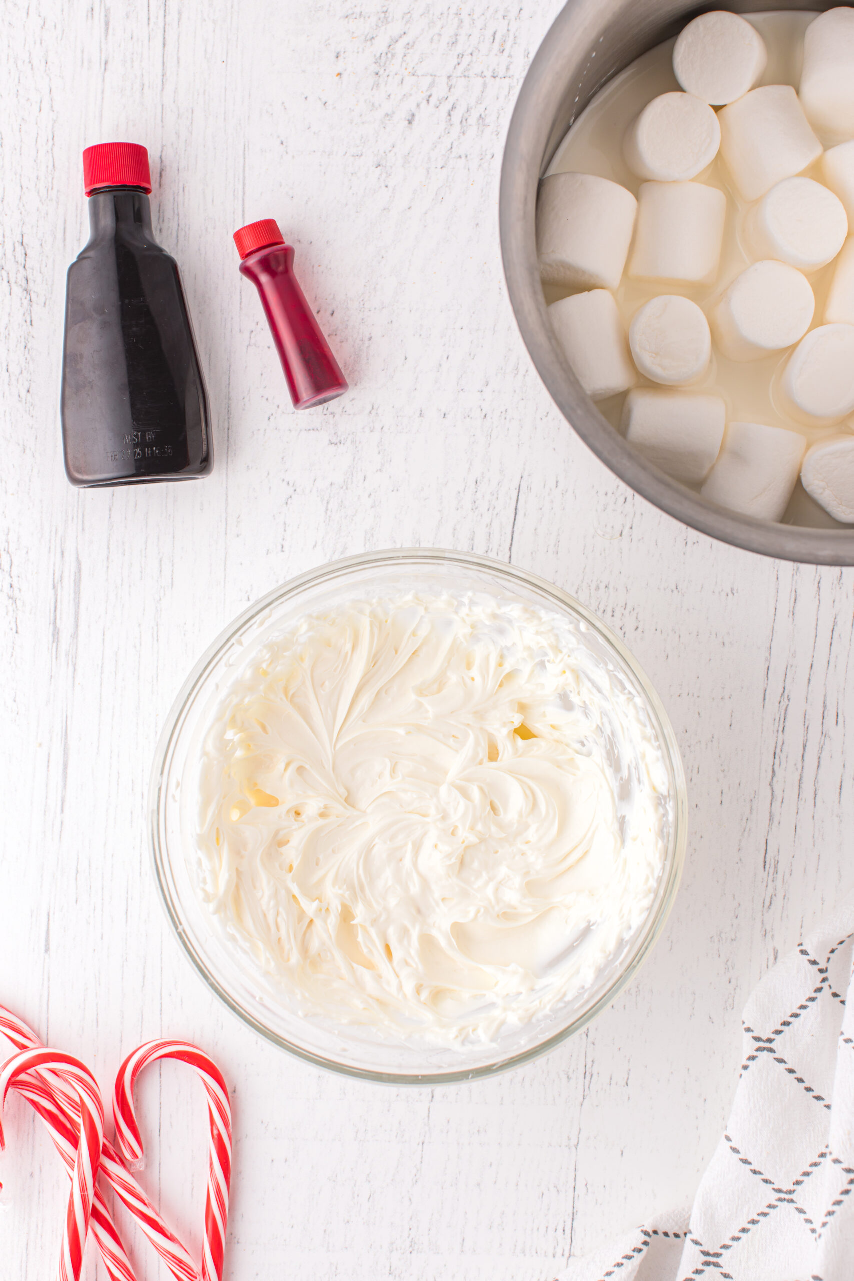 whipped cream cheese in a bowl with small bottle of peppermint extract and red food coloring next to it. bowl of large marshmallows in upper right corner candy canes in bottom left corner.