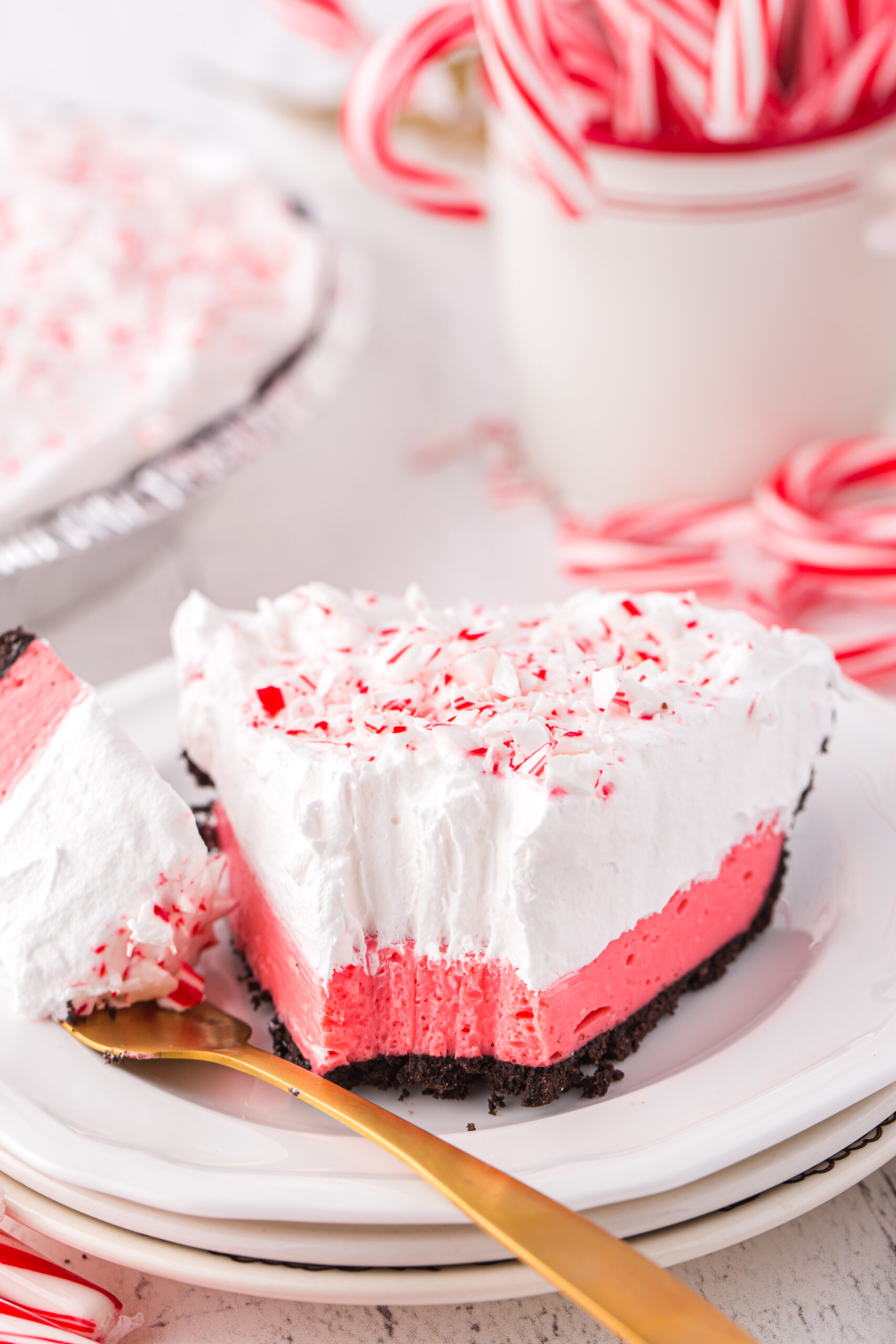 slice of pretty layered peppermint pie on a small white plate with a bite taken out of it with a fork. Gold fork with pie on it resting next to the pie on the plate. Whole pie teased in the background.
