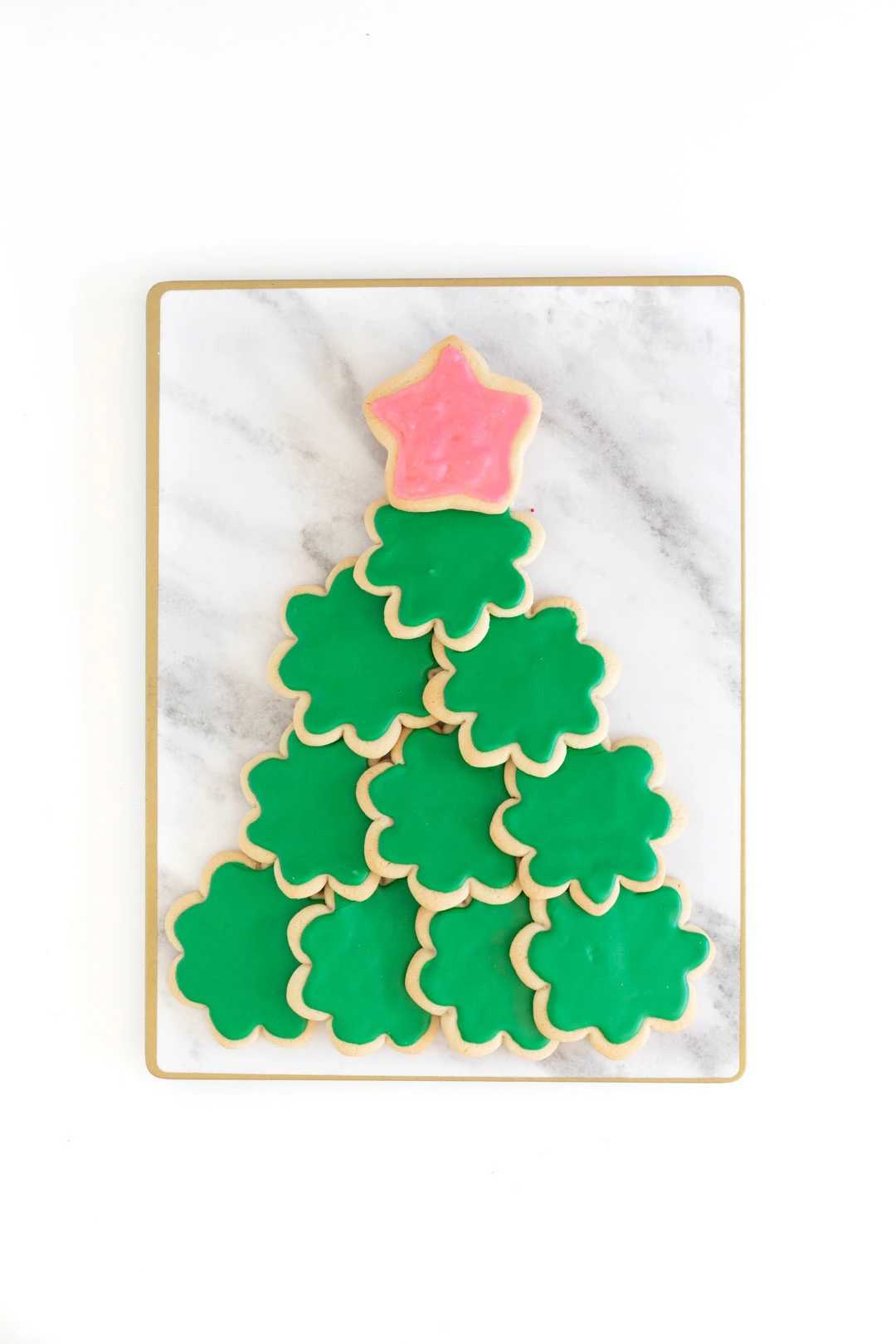 a cookie christmas tree being prepared on a marble colored cutting board. green frosted sugar cookies for the base and pink frosted star for the top