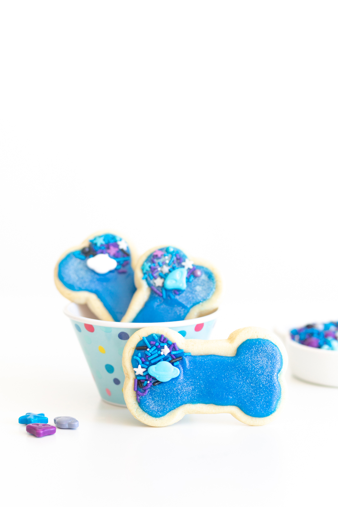 galaxy dog bone cookies with bright blue icing and alien sprinkles. Cookies placed in blue polkadot bowl with one leaning outside the bowl